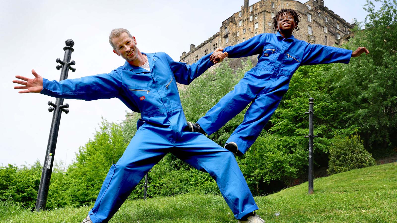 A man and a boy in bright blue jumpsuits in a garden. They are dancing joyfully with each other, striking a pose with the boy standing on the mans legs as they stay connected by holding hands.