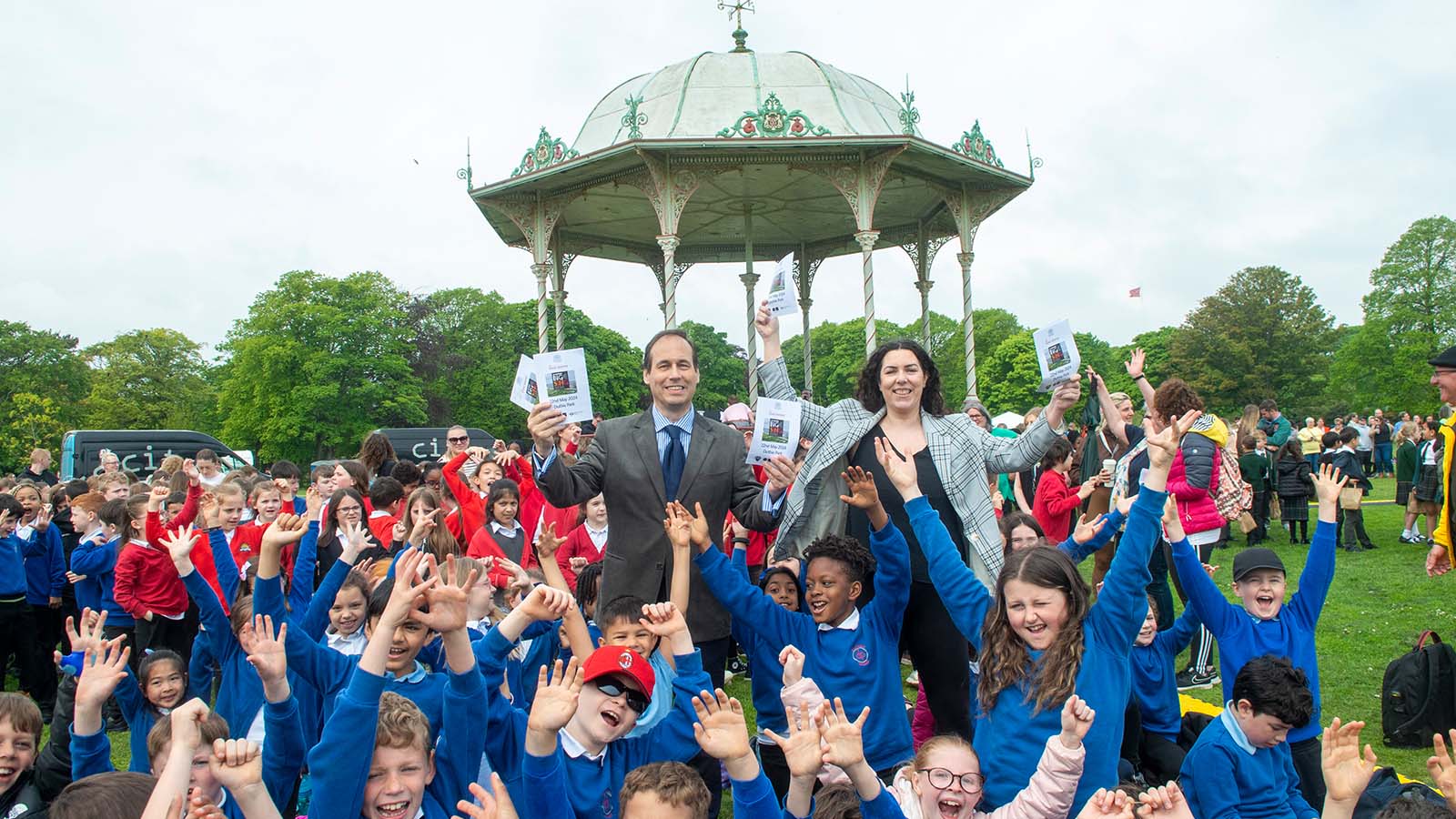 Primary school pupils in bright blue and red school uniforms crowd together in a park, their arms raised in celebration, in the centre, two adults holding the programmes for The Big Sing, in the background, the gazebo where the band performed
