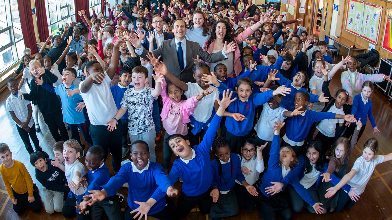 A large group of smiling children in a school gymnasium, their arms raised as they sing. Three adults stand at the centre, smiling and singing alongside them.