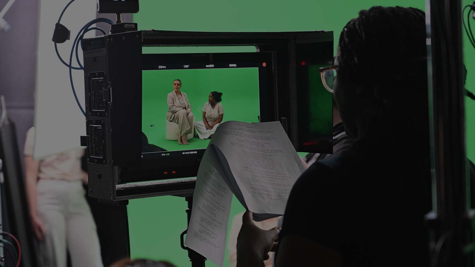 A film crew captures a green screen scene with two actors seated, while a crew member reviews a script in the foreground.