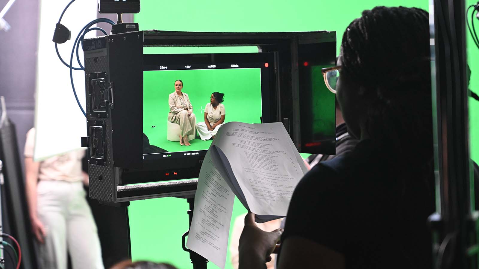A film crew captures a green screen scene with two actors seated, while a crew member reviews a script in the foreground.