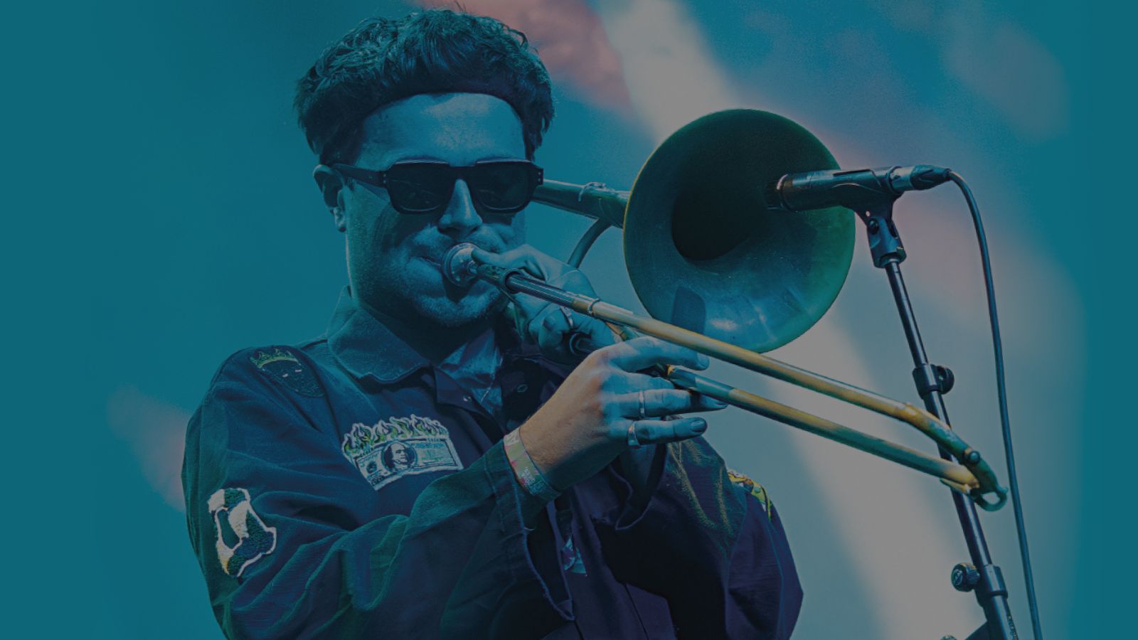 A musician plays a brass trombone on stage, wearing a dark jacket with colorful patches against a vibrant blue background.