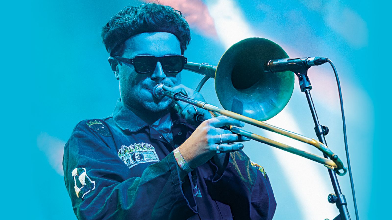 A musician plays a brass trombone on stage, wearing a dark jacket with colorful patches against a vibrant blue background.