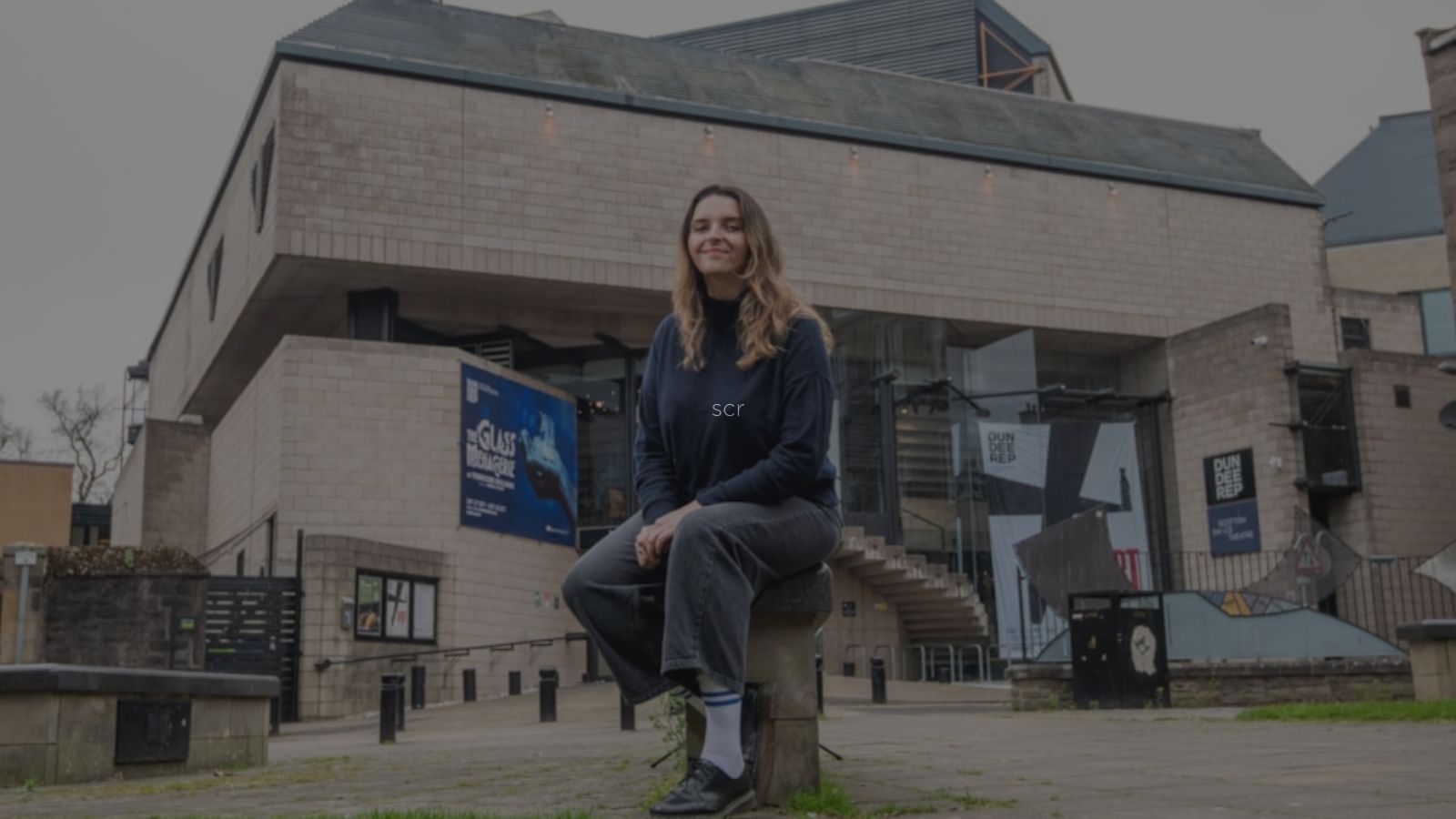 A person sits on a stone in front of a modern building with large windows and promotional banners. The atmosphere is overcast and urban.