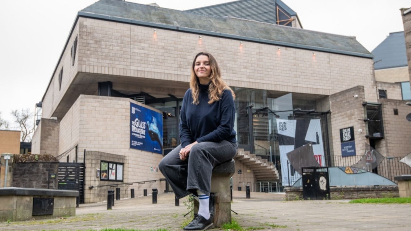 A person sits on a stone in front of a modern building with large windows and promotional banners. The atmosphere is overcast and urban.