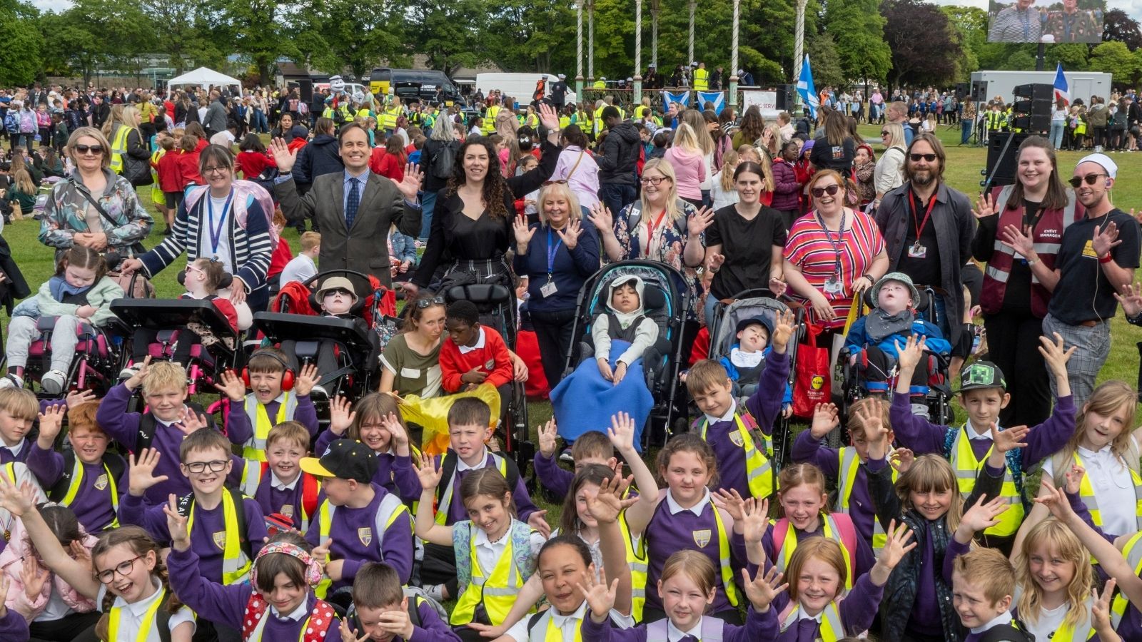 A lively outdoor gathering featuring children in bright vests and adults, all smiling and waving in front of a decorative bandstand.