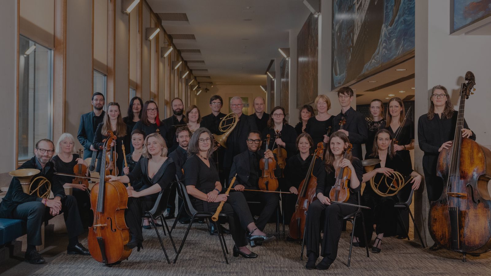 A diverse orchestra posed indoors, dressed in black, with various instruments including strings and brass, against a bright, modern backdrop.