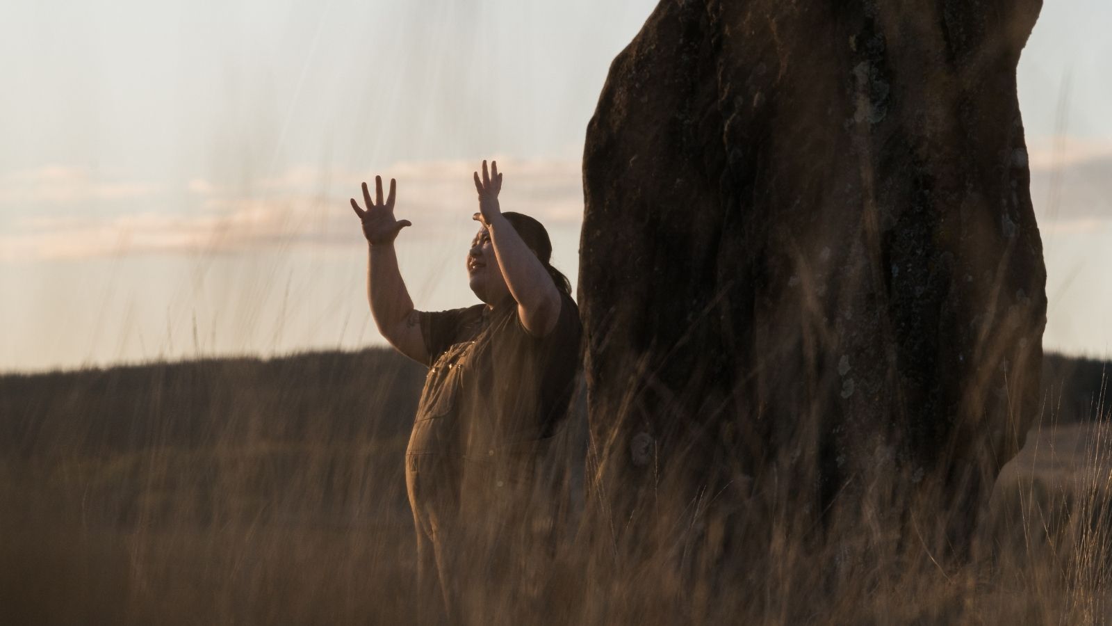A person stands beside a large rock, raising both hands against a soft sunset backdrop with tall grass in the foreground.