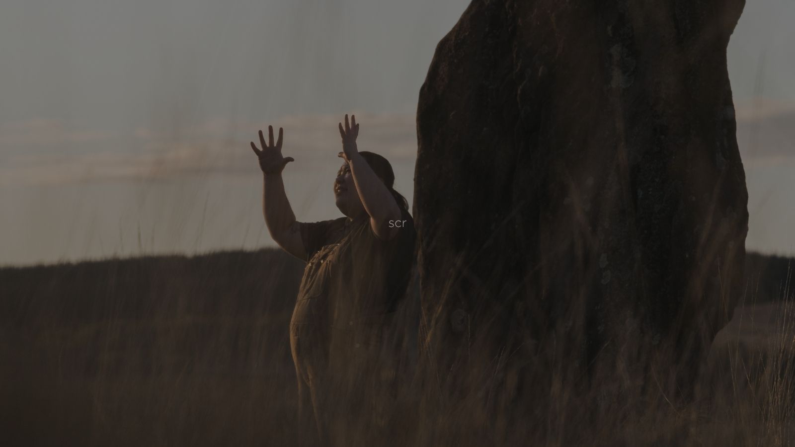 A person stands beside a large rock, raising both hands against a soft sunset backdrop with tall grass in the foreground.