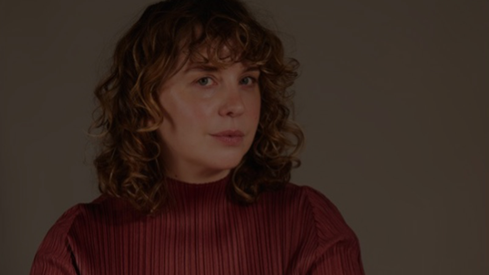A person with curly hair wearing a ribbed maroon sweater stands against a soft, neutral background, exuding a calm demeanor.
