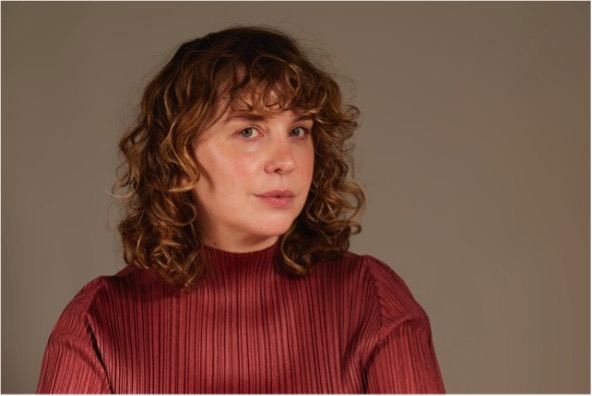 A person with curly hair wearing a ribbed maroon sweater stands against a soft, neutral background, exuding a calm demeanor.