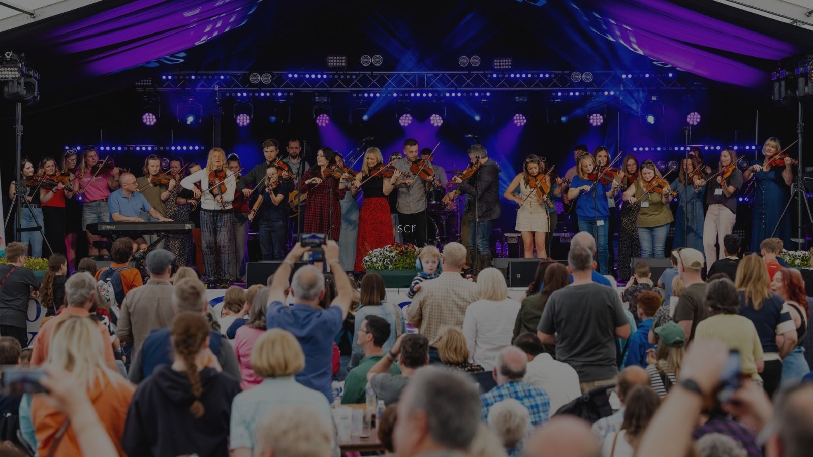 A lively outdoor concert performance featuring a diverse group of violinists on stage, with an engaged audience in front.