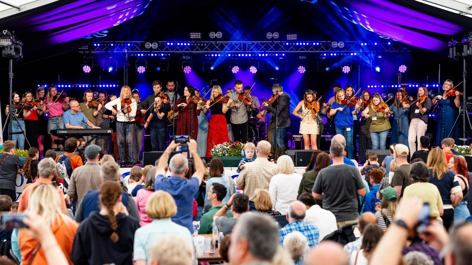 A lively outdoor concert performance featuring a diverse group of violinists on stage, with an engaged audience in front.