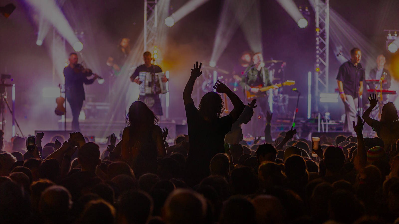 A vibrant concert scene with an energetic crowd, hands raised, and a band performing on stage surrounded by colourful lights.