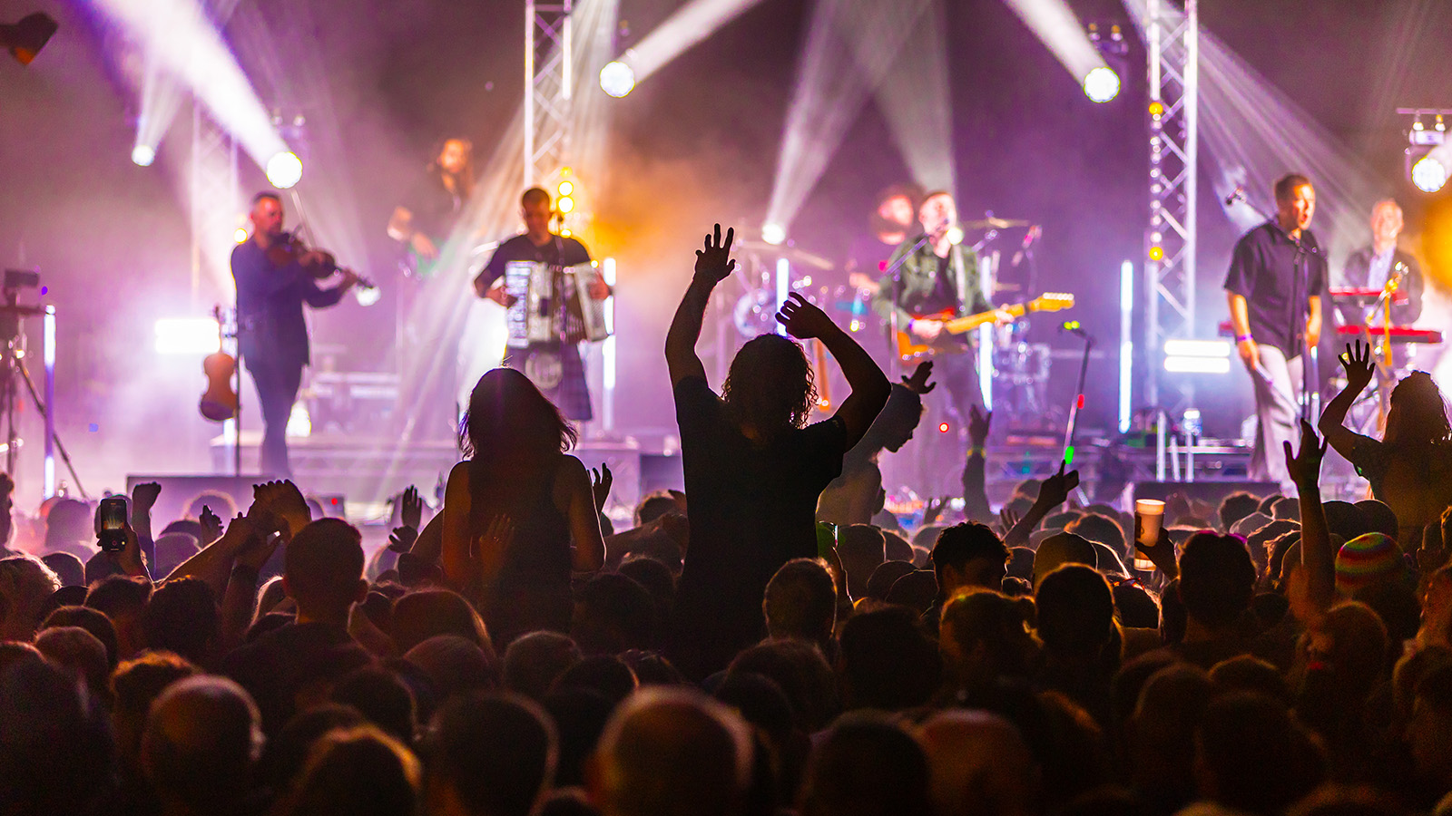 A vibrant concert scene with an energetic crowd, hands raised, and a band performing on stage surrounded by colourful lights.