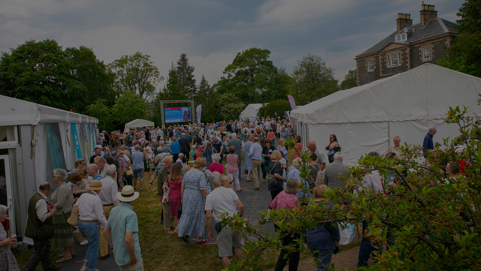 A large crowd gathered outdoors at an event, with white tents, greenery, and a large screen displaying information in the background.