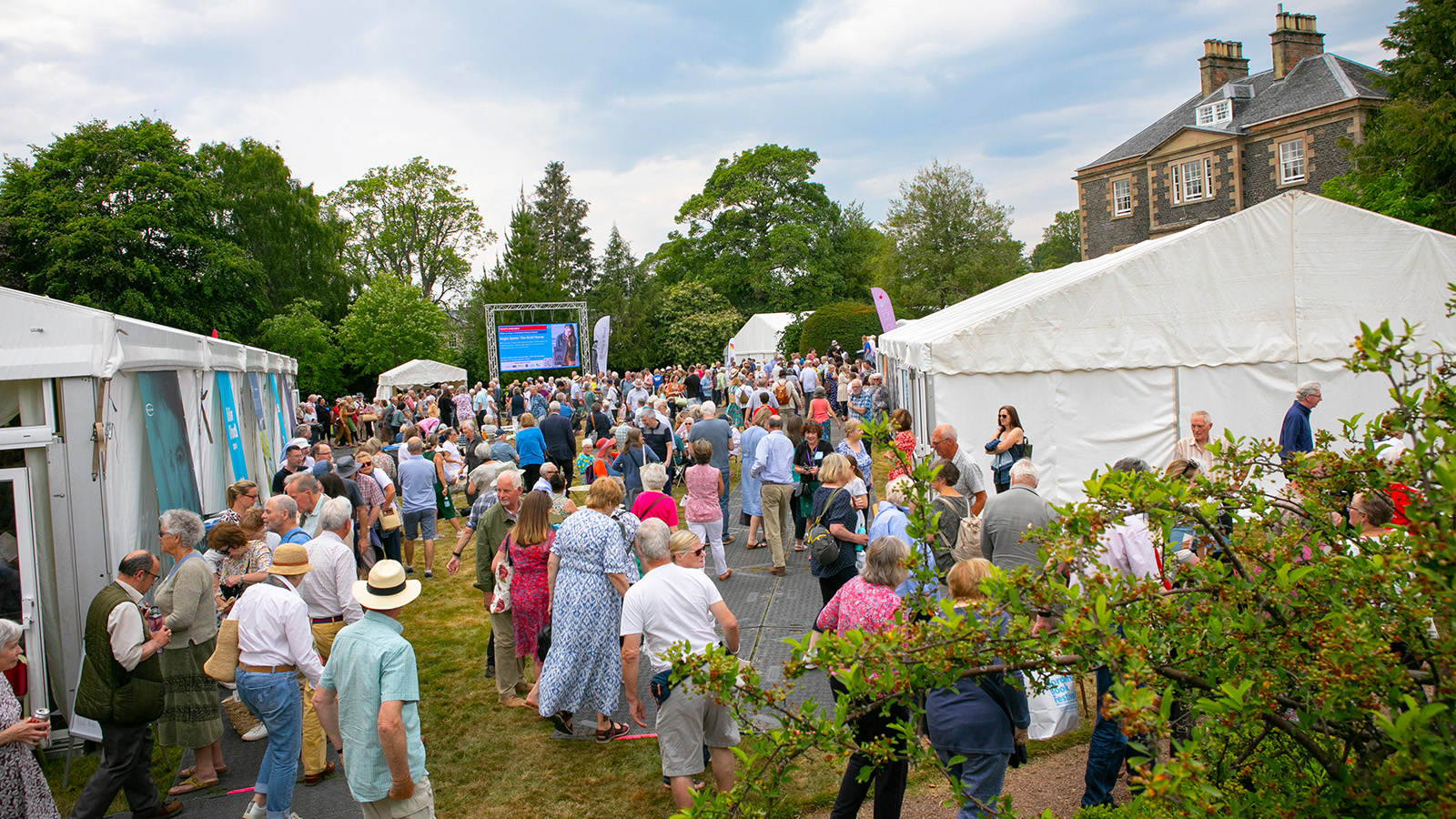 A large crowd gathered outdoors at an event, with white tents, greenery, and a large screen displaying information in the background.