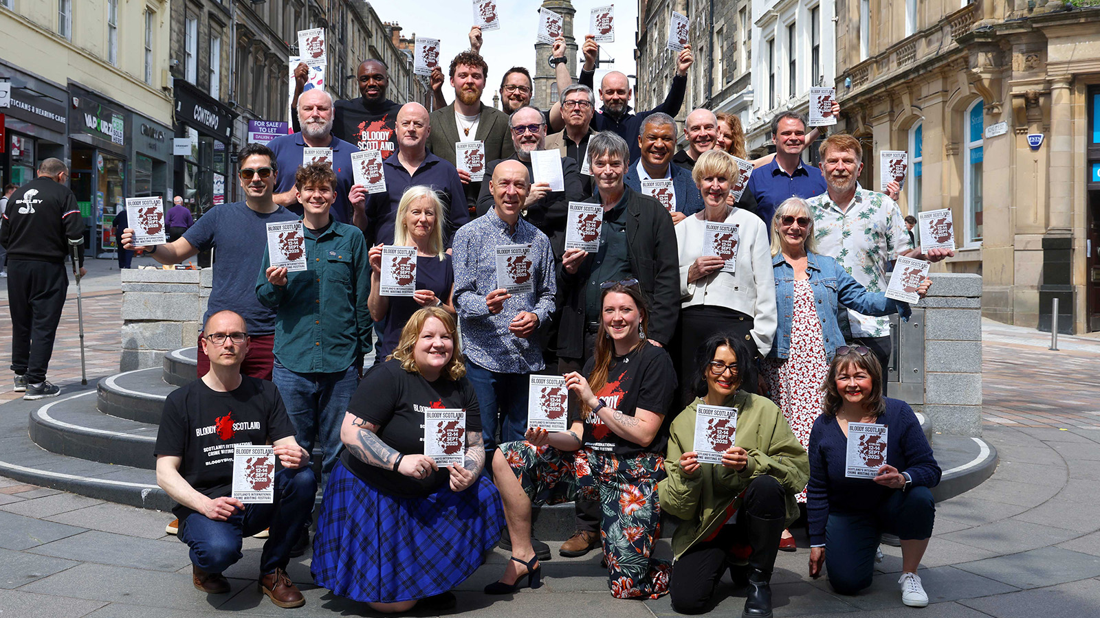 A diverse group of individuals gathers in a city square, holding event flyers for "Bloody Scotland." Buildings and shops line the background.