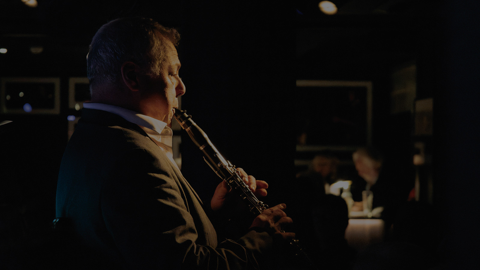 A musician in a suit plays the flute on stage in a dimly lit venue, with patrons enjoying the performance in the background.