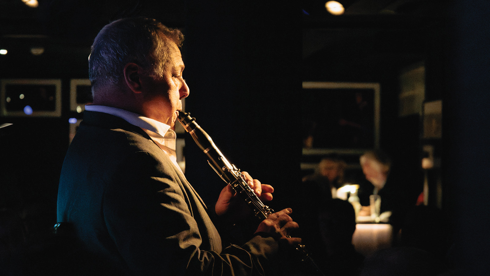 A musician in a suit plays the flute on stage in a dimly lit venue, with patrons enjoying the performance in the background.