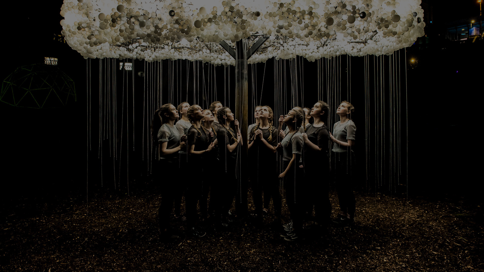 A group of individuals stands beneath a large, illuminated cloud sculpture, surrounded by atmospheric lighting and a dark, forested setting.