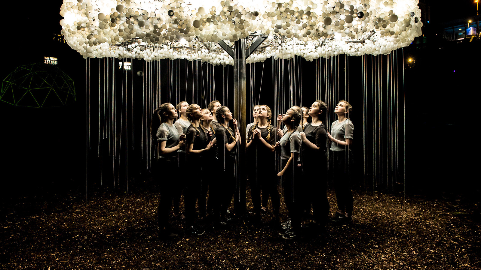 A group of individuals stands beneath a large, illuminated cloud sculpture, surrounded by atmospheric lighting and a dark, forested setting.