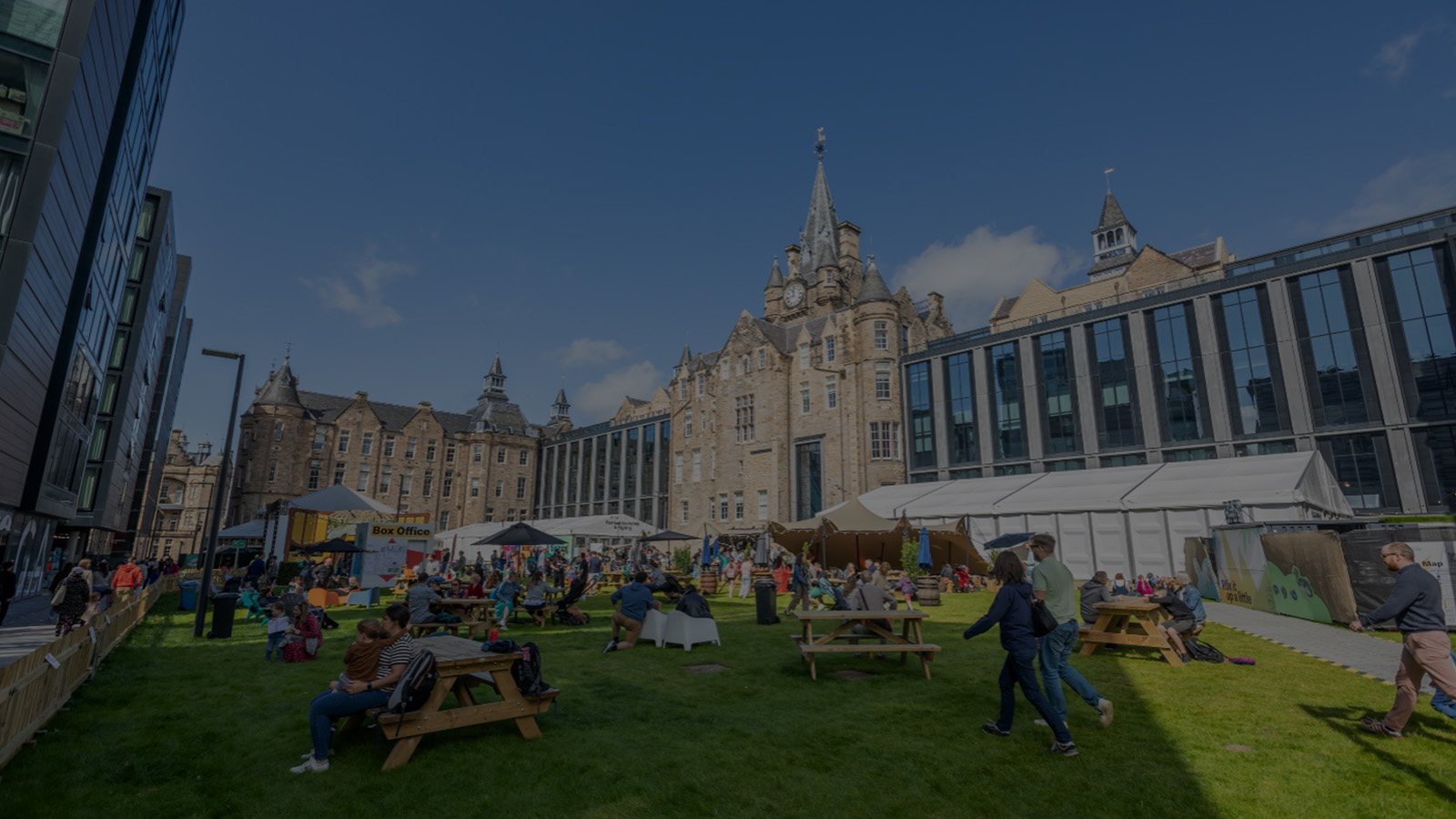 A bustling outdoor market scene in a city park, surrounded by historic and modern buildings, with people enjoying food and activities.