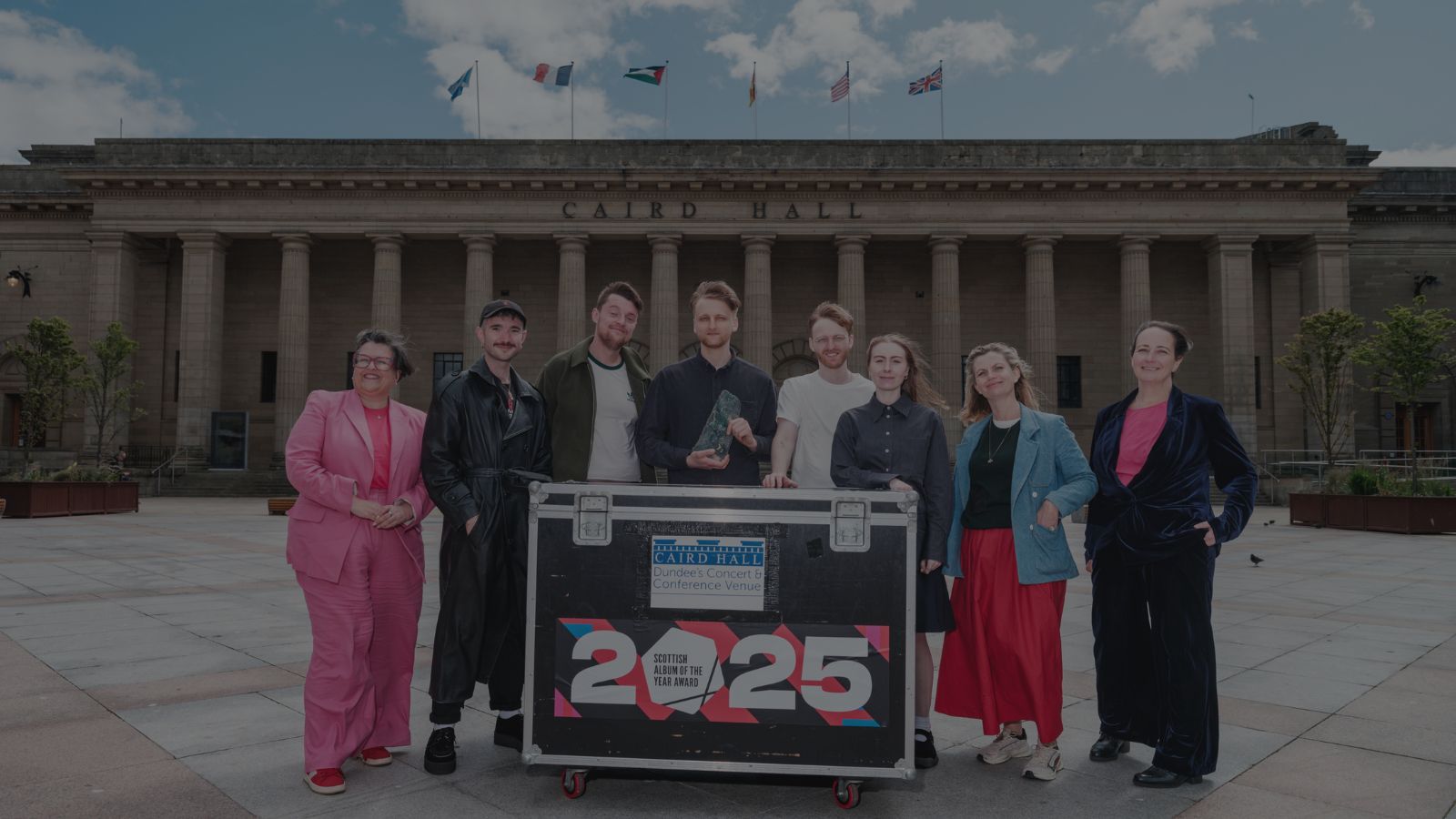 A group of people stands in front of a large stone pillared building, the Caird Hall, with a large crate, which reads Caird Hall, Scottish Album of the Year Award 2025.