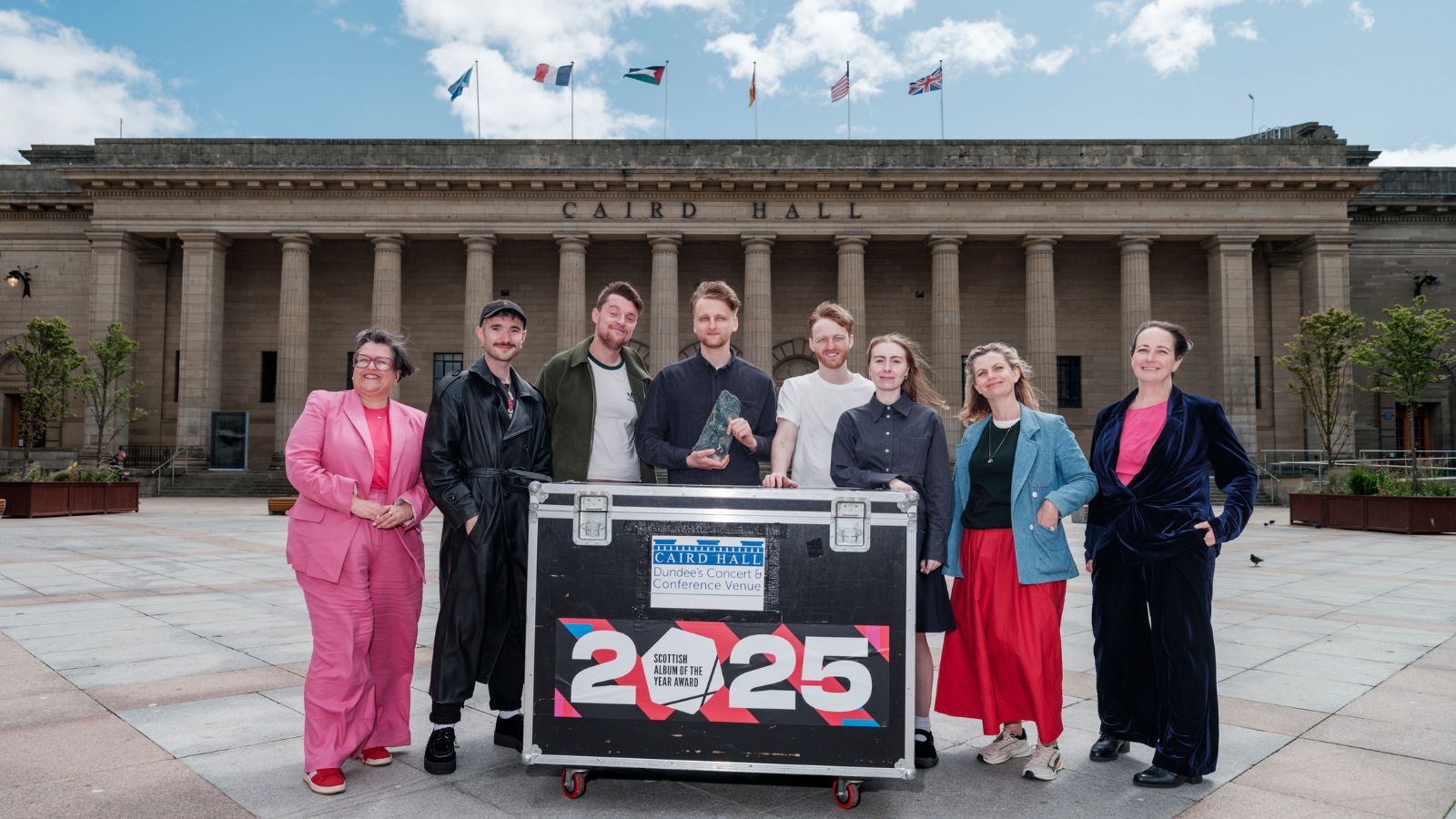 A group of people stands in front of a large stone pillared building, the Caird Hall, with a large crate, which reads Caird Hall, Scottish Album of the Year Award 2025.