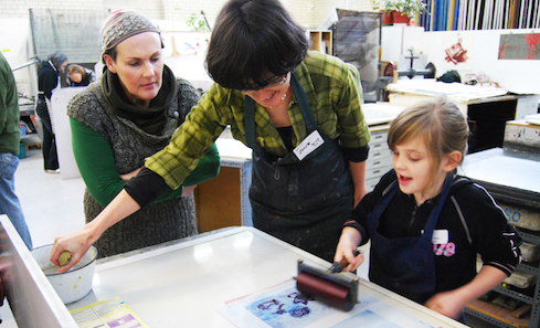 A woman stands in front of print making materials teaching a child how to coat a stencil in ink
