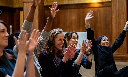 A group of people in a wood panelled room have their hands up, some clap, they all look excited