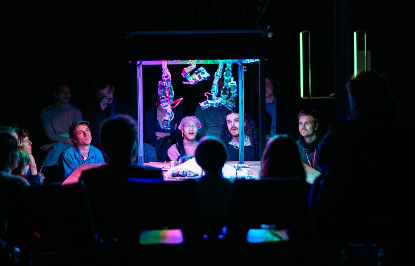 Performers and audience members sit at a table looking at a variety of brightly lit objects as part of the Simple Machines show